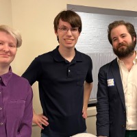 Three people pose together with hands on hips and hands on a chair in front of him.